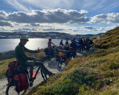 Lærer og elever på skykler, på en sti. Innsjø og fjell i bakgrunnen. 