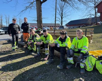 Anleggselever sitter på rekke. Ved siden av står to lærere.