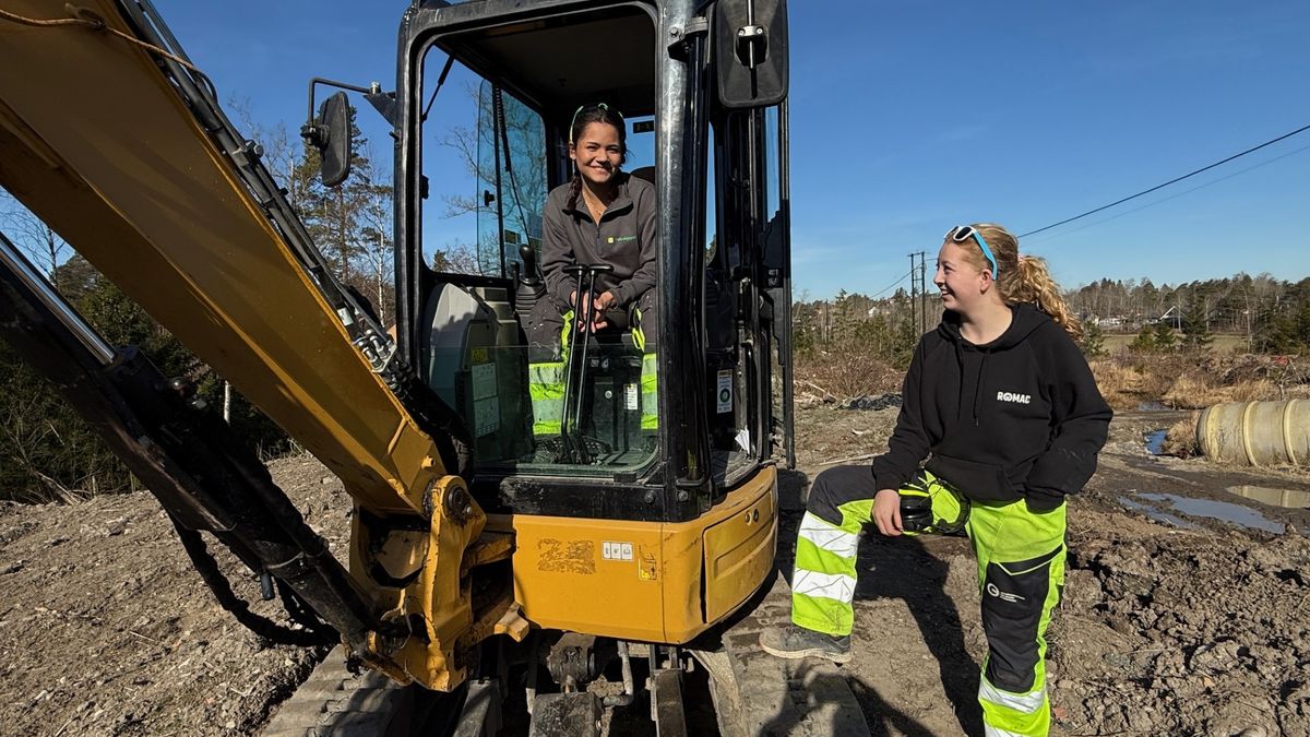 En elevjente sitter i i en gravemaskin på et anleggsområde. En medelev står ved siden av.